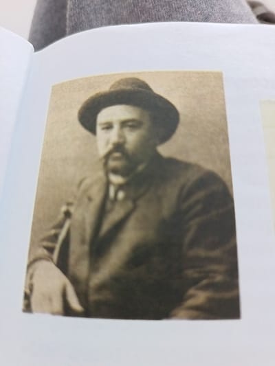 A sepia-toned photograph depicts a man wearing a dark hat and a suit. He has a thick mustache and a neatly trimmed beard. His hand rests on something out of frame. The picture appears to be from an old book or document, evidenced by the surrounding white pages. The man is centered, posing from the waist up, with a slight turn towards the viewer. The image has a vintage or historical feel.