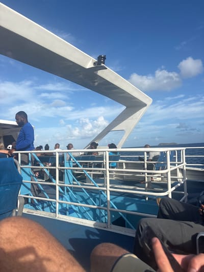 The scene depicts individuals aboard a passenger vessel, likely a ferry or catamaran, traveling at sea in the vicinity of Spanish Town, British Virgin Islands. The perspective is from a low angle, looking forward from a seated position.
In the immediate foreground, the bare, tanned, and hairy legs of one individual wearing dark shorts are visible on the left, alongside the legs of another person in dark pants on the right, whose right hand holds a mobile phone.
The midground features the vessel's deck and passenger area. A white, angular structural support beam extends diagonally across the upper portion of the frame, with a black cylindrical object mounted on its underside. Below this, a series of white metal railings with blue accents delineate seating sections. A standing male with a dark complexion, wearing a long-sleeved blue shirt and dark trousers, is prominent on the left, positioned near a cabin entrance. He faces towards the left, partially obscured. Behind the railings, at least six to eight other passengers, primarily adults with dark complexions, are visible seated in blue chairs. Their actions appear passive, consistent with traveling. One individual on the far right, possibly a child, wears a light blue top.
The background shows the open ocean with moderate choppiness. A small, low-lying green island is visible in the distance on the horizon to the far right. The sky is bright blue with scattered white cumulus clouds, indicating fair weather conditions.