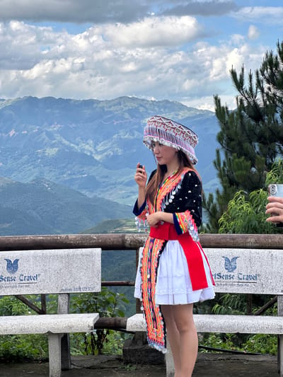 A woman in traditional attire stands outdoors at a scenic viewpoint in Tu Le, Vietnam, with a mountainous landscape in the background. The woman, positioned slightly right of center, faces left with her head tilted slightly up. She wears an elaborate headpiece featuring intricate white, red, and black patterns with numerous dangling silver-colored discs. Her long-sleeved black top is adorned with colorful embroidery in red, yellow, and blue, and also features silver-colored disc embellishments. She has a bright red sash around her waist and a white, pleated skirt. A long, colorful embroidered panel, matching the style of her top, hangs down her left side. In her right hand (viewer's left), she holds a small, dark, cylindrical object. To her right (viewer's right), a hand holding a silver-colored smartphone with a multi-lens camera array is visible, pointing towards her, indicating she is being photographed.
In the immediate foreground, a rustic metal railing separates the viewer from the scene. Behind the railing are two speckled gray granite benches. The bench on the left displays the logo and text "Sense Travel®" along with partially visible Vietnamese text "lich voi niem dam me" and a hotline number "0916 172 338". The bench on the right also shows the "Sense Travel®" branding.
The background consists of extensive, verdant mountains covered in dense foliage, with visible terraced fields and scattered structures on the slopes. The sky above is partly cloudy, featuring large white and gray cumulus clouds with patches of blue sky. A tall pine tree with long needles is partially visible on the far right. Green shrubs and plants grow behind the benches and railing.