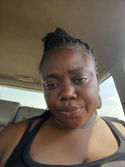 This media file shows a close-up, low-angle selfie of a Black person with dark skin and braided hair. The person is wearing a black tank top and a nose ring. They are looking at the camera with a slight smile, and their head is tilted. The background appears to be the interior of a car, with a beige headliner and seat visible. The image is taken during the day, likely with natural light coming from the sky through a sunroof or window, suggesting it might be an overcast day. The setting is likely within Sabojibi, Nigeria.
