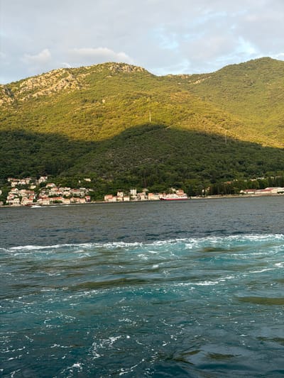 This image depicts a body of water in front of a coastal town nestled at the foot of a lush, green mountain. The town consists of buildings with white walls and red roofs, clustered together along the shoreline. The mountain behind the town is densely covered with trees, with rocky outcrops visible in some areas. The sky above is partly cloudy, with sunlight casting a warm glow on the mountain and the town. The water in the foreground is choppy, with white foam indicating movement, possibly from a boat or a strong current. The scene appears to be a peaceful, scenic view of a coastal settlement in Đurići, Montenegro.