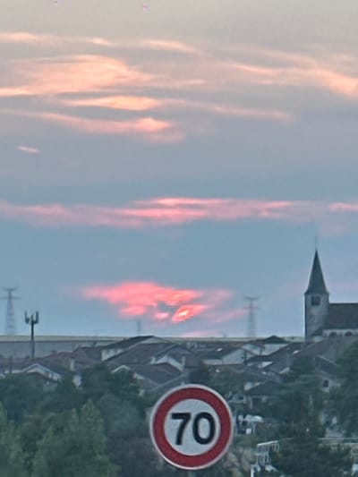 The image depicts a scenic view in Pagny-sur-Meuse, France, with a sunset casting a vibrant pink glow across the sky. In the foreground, a red and white circular speed limit sign displays the number "70". Behind the sign, a dense cluster of trees partially obscures a collection of houses with reddish-brown roofs. A church steeple, characteristic of French villages, is visible in the middle ground, to the right of the frame. In the distance, power pylons stand tall against the twilight sky, and a faint line of buildings can be discerned. The overall atmosphere is serene and picturesque, capturing the tranquil beauty of a French countryside sunset.