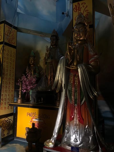 The image shows a dimly lit interior, likely a temple or shrine, in Nam Ban, Vietnam. The focal point is a large, golden statue of a deity with intricate details, positioned prominently in the foreground. The deity is adorned with elaborate jewelry and flowing robes, and its hands are raised in a gesture of blessing or greeting.
To the left of the main statue, a smaller, green-hued statue is visible, also with hands raised. A vase filled with pink flowers sits in front of this smaller statue. Behind them, a tall, yellow pillar is decorated with what appear to be religious symbols or script in red.
Further in the background, another, taller statue can be seen, partially obscured. The environment is characterized by a blue, possibly painted, ceiling and a yellow, rectangular panel with red lettering. The overall atmosphere is serene and spiritual, emphasizing the religious nature of the location. The lighting is dramatic, with shadows playing a significant role in defining the forms and creating depth.