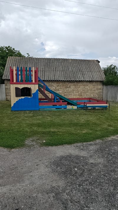 In Revne, Ukraine, a colorful, handmade wooden playground structure is situated on a grassy area in front of a stone building with a corrugated roof. The playground features a multi-level tower with a red and blue fence, a slide with blue and green ramps, and red railings. To the right of the slide, a simple wooden bench with black metal legs is placed on the grass. A gravel path runs along the foreground of the image. The sky above is overcast with scattered clouds.