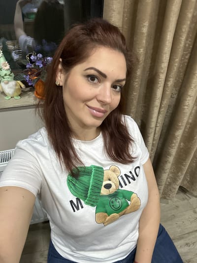 A woman with reddish-brown hair and fair skin is centered in the image, wearing a white t-shirt with a cartoon bear graphic in green. She is wearing gold earrings. The background features a neutral-toned curtain on the left and what appears to be blue denim on the right. A decorative floral arrangement is visible to the left. The location and city of Lehchevo, Bulgaria, are not discernible from the provided image.