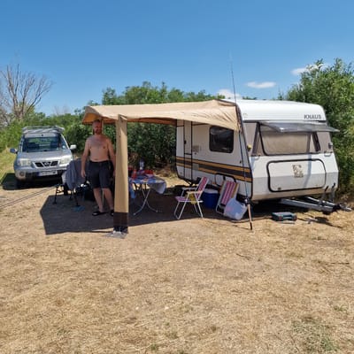 A bare-chested man with short hair and a beard stands in front of a caravan at a campsite. He is wearing dark shorts and dark sandals. The caravan is white with yellow and grey stripes and the KNAUS logo. An awning extends from the caravan, providing shade over a table set with plates, food, and drinks. Folding chairs are set up around the table. To the left of the caravan is a silver Nissan X-TRAIL SUV with a license plate reading "EH 0577 MK". The ground is covered in dry grass and dirt. In the background, there are green bushes and trees, and a bright blue sky with scattered clouds. The scene appears to be set in the city of Gorni Dabnik, Bulgaria, suggesting a recreational camping activity.