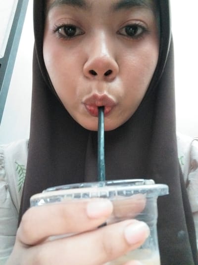 The image is a close-up, head-and-shoulders shot of a person wearing a brown hijab, holding a clear plastic cup with a straw. The person's face is partially obscured by the cup and straw as they appear to be drinking. Their lips are pursed around the straw, and they are looking directly at the camera. The background is mostly white with some light gray. The person is wearing a light-colored garment with a green leaf pattern visible on the left side of the image. The scene is indoors, likely a casual setting. The specific location in Tibojong, Indonesia cannot be determined from the visual information provided.