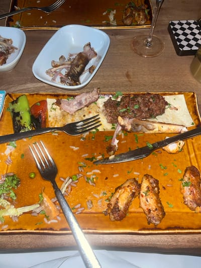 This image presents a forensic view of a dining table in Stevenage, United Kingdom, immediately following a meal. In the foreground, a rectangular, dark orange ceramic plate is largely cleared but contains scattered remnants: three partially consumed chicken wing sections, grains of white and brown rice, green peas, and finely chopped green herbs. A grilled green pepper and a grilled red tomato half are present on the left side of the plate. Additional remnants include a portion of flatbread, a minced meat patty, and several bone fragments, including a larger rib-like bone. A silver-colored fork with a textured handle rests diagonally on the plate's left, and a matching knife is positioned horizontally on the right.
In the mid-ground, a white, rectangular ceramic bowl, tilted slightly, is filled with numerous clean bone fragments, indicating a discarded collection from a previous course. A second, similar white bowl containing bones is partially visible to the left. The table surface is a light-colored wood. To the right, a stemmed wine glass, containing minimal liquid, stands next to a black and white checkered coaster. A golden-colored cylindrical object is partially visible behind the coaster. In the blurred background, two additional, mostly cleared rectangular dark brown plates are visible with scattered food debris and cutlery, confirming a multi-person dining event. No individuals are present in the scene.
