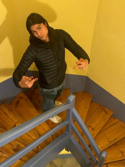An individual with long, dark hair stands on a wooden spiral staircase in Roanne, France. The person wears a black puffer jacket, blue jeans, and white sneakers. Both hands are raised to chest level, displaying red-painted fingernails, with the index and middle fingers extended on each hand. The staircase features light brown wooden treads and a blue-grey painted handrail and balustrade. The walls are painted yellow with a matching blue-grey stripe at the base. A shadow is cast on the upper left yellow wall.