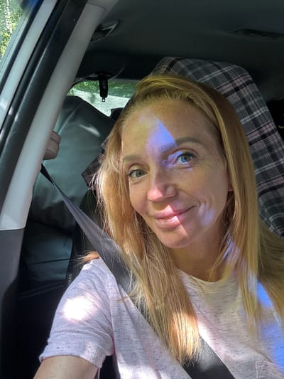 An outdoor, close-up, eye-level, selfie photograph shows a fair-skinned blonde woman with blue eyes and freckles, smiling in a car. The woman is wearing a light pink t-shirt and a seatbelt. A blue light reflects on her face. Behind her, a plaid pattern is visible, likely from a car seat cover or blanket. The woman is in the passenger seat of the car, and the background shows greenery outside the window. The context of Portovoe, Russia is not discernible from the image.