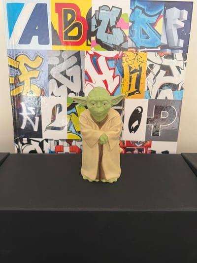 The image shows a figurine of Yoda from Star Wars standing on a black surface. Behind the figurine is a wall covered with graffiti art, featuring various letters and designs in different colors. The graffiti appears to be a mural or a display of stylized lettering. The lighting is neutral, suggesting an indoor environment. There is no visible indication of the location being in Malmö, Sweden.