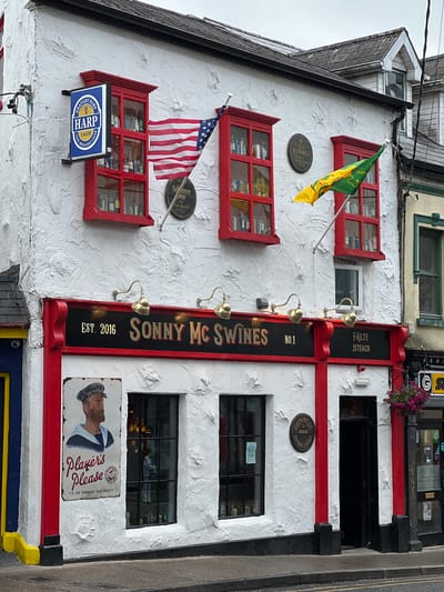 The image shows the exterior of "Sonny Mc Swines" pub, located in the city of Donegal, Ireland. The pub has a white, textured facade with vibrant red trim around windows and doorways. Above the entrance, a prominent sign reads "EST. 2016 SONNY MC SWINES NO.1 FÁILTE ÍSTEACH".

Several flags are visible: an American flag is mounted to the building, and a green and yellow flag, possibly representing Ireland, is flying from a pole. Various signs and logos adorn the building, including a "HARP LAGER" sign, an "OLD BUSHMILLS" plaque, and a "GUINNESS DUBLIN EXTRA STOUT" plaque. A vintage advertisement featuring a sailor with the text "Player's Please 'IT'S THE TOBACCO THAT COUNTS'" is displayed near the entrance.

The windows of the pub appear to be displaying bottles, likely alcoholic beverages. The building has multiple stories, with windows on the upper floors also visible. The street in front of the pub is paved, and a lamppost with hanging flowers stands to the right. The overall scene suggests a lively and traditional Irish pub setting.