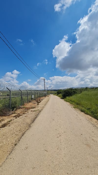 The image shows a rural scene in Hof Ashkelon Regional Council, Israel. A dirt road stretches into the distance, bordered on one side by green vegetation and on the other by a fence topped with barbed wire. Utility poles run parallel to the fence. The sky is blue with white, puffy clouds.