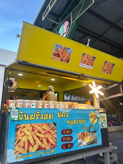 A food cart selling French fries is parked on a street in Tha Khlong Town Municipality, Thailand. The cart is yellow with a blue tarp displaying the menu. A woman is seen working at the cart, serving customers. The weather is overcast, and it appears to be late afternoon or early evening. The food cart is decorated with pictures of French fries and various dipping sauces. There are also QR codes for payment. The cart has a selection of seasonings displayed in clear containers. The street is paved, and there are trees and buildings visible in the background. A motorcycle is parked nearby, suggesting the area is a public space. The overall scene depicts a typical street food vendor operation in Thailand.
