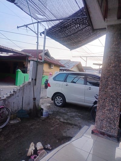 A white van is parked in front of a building. A person on a motorcycle is passing by. A bicycle is leaning against a wall. There are brooms and a rake on the ground, as well as a bucket. Several pairs of shoes are scattered on the ground. The scene appears to be in a residential area, possibly in Indonesia, given the architecture and the presence of a motorcycle. The sky is clear, suggesting it is daytime, likely late afternoon or early morning due to the soft light. The overall impression is one of a quiet, everyday scene.