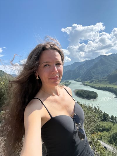 A woman with long, wavy brown hair takes a selfie outdoors with a scenic mountain and river landscape in the background. She is wearing a black tank top with thin straps and a delicate gold necklace. Sunglasses are clipped to the front of her top. The woman is smiling slightly as she looks at the camera. Her hair is blowing in the wind to her left. The background shows a turquoise river winding through a valley with lush green trees and tall mountains under a bright blue sky with scattered white clouds. A small island covered in trees is visible in the river. The image appears to have been taken from a high vantage point, likely overlooking the Chemal River in the Altai Republic, Russia, potentially near the confluence with the Katun River, a common tourist area for its natural beauty.
