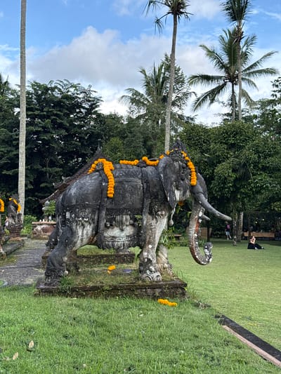 The image shows an elephant statue in a park setting in Carangsari, Indonesia. The statue is adorned with orange garlands. Palm trees are visible in the background. In the upper right, people can be seen relaxing on the grass. The statue sits on a stone base surrounded by grass. The sky is partially cloudy.