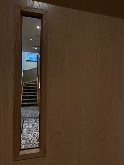 A wooden door features a long, rectangular peephole. Through the opening, a glimpse into an interior is revealed. The view extends from a brightly lit, possibly windowed room, transitioning to a staircase with dark carpeting, and ending with a wall covered in decorative wallpaper. The peephole's wooden frame creates a contrasting border to the scene it contains.
