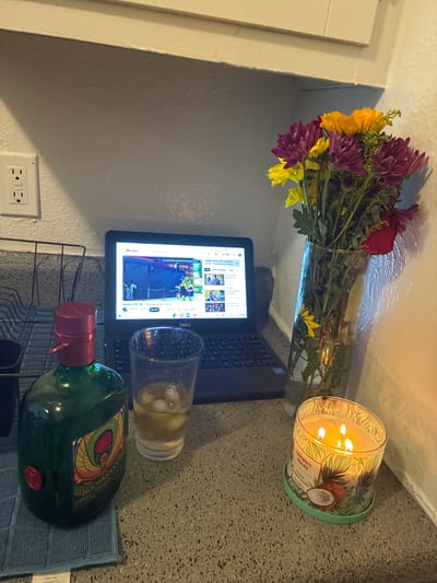 This is a still image of a domestic scene, likely a kitchen counter or desk area. The scene is illuminated by artificial light, possibly from a candle and the laptop screen, suggesting it might be evening or nighttime.

The primary subjects include a bouquet of purple and yellow flowers in a clear glass vase, a lit scented candle with a tropical design and the text "COCONUT COAST" and "3-wick Scented candle", a laptop displaying a YouTube video interface with the text "Siempre StRf The Rojas View Oncia" visible, and a glass containing a yellowish liquid with ice. A green bottle of Buchanans Deluxe whiskey is also prominently featured, resting on a blue cloth. An electrical outlet is visible on a light-colored wall in the background.

The scene appears to be set indoors, given the presence of the laptop, candle, and electrical outlet. The overall mood is relaxed and perhaps solitary, with the combination of flowers, a candle, and a drink suggesting a moment of personal downtime. The specific location "Balch Springs, United States of America" provides geographical context. There are no people visible in the image.