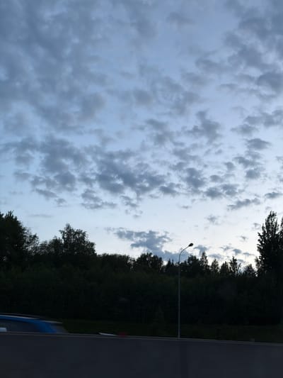 The image presents an outdoor scene dominated by a cloudy sky and a line of dark trees. The perspective appears to be from a slightly elevated position or looking upwards, showing a vast expanse of sky.
The upper two-thirds of the frame are filled with a complex pattern of grey, lumpy clouds, suggesting a heavily overcast or impending storm condition. These clouds exhibit varied textures and densities, with some areas appearing darker and more voluminous, while others reveal lighter, brighter patches of pale blue-grey sky peeking through, particularly towards the right side of the image. The cloud formations are somewhat chaotic but largely cover the firmament.
Across the lower third of the image, a dense and dark line of trees forms a natural boundary, appearing largely as a silhouette against the brighter sky. These trees are heavily foliated, indicating a possible summer or late spring setting. A single, tall street light with a distinctive curved arm rises from among these trees, extending upwards into the cloudy sky, positioned slightly to the right of the center. In the very bottom right corner, a dark grey structure, possibly a road barrier or wall, is visible, alongside a small, indistinct blue section of what appears to be a vehicle, likely a car.
The overall lighting suggests it is either late afternoon, early evening, or a day with very heavy cloud cover, as there is no direct sunlight visible and the landscape elements are silhouetted. No specific activity is taking place. There is no visible text. The location context is Kerro Massiv, Russia.