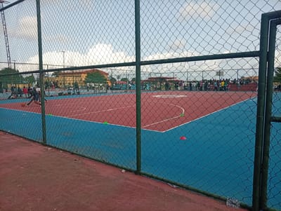 The image shows an outdoor basketball court located in Ijanikin, Nigeria, enclosed by a chain-link fence. The court surface is two-toned, featuring a red area transitioning into a blue area. Several orange and green cones are scattered on the court. A group of people is visible behind the fence, appearing to be spectators or participants gathered on the sidelines. In the background, buildings and trees are partially visible under a cloudy sky. A person is dribbling a basketball on the court.