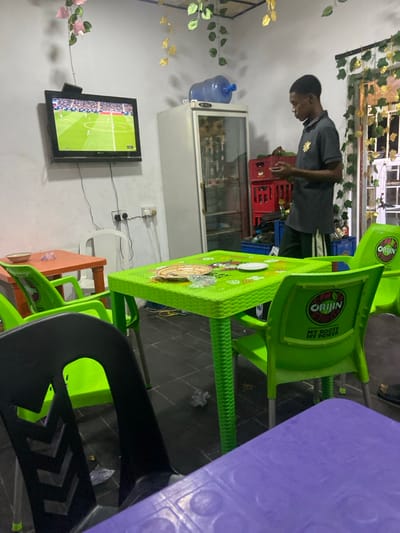 A room with green plastic chairs and tables, including purple and black ones, is visible. A man in a grey shirt stands near a refrigerator. A TV on the wall shows a soccer match. The room is decorated with artificial flowers. Crates are stacked near the refrigerator. A water dispenser is also mounted on the wall.