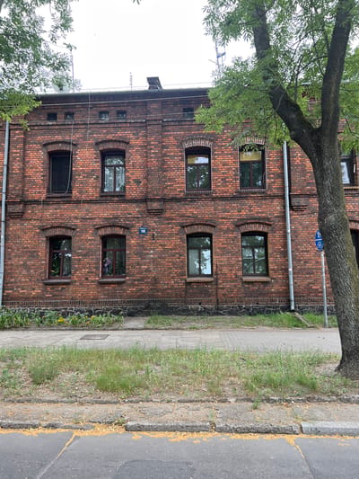 This is a medium-shot, eye-level, exterior, daytime photograph of the brick facade of a multi-story building. The building has at least two floors visible, with several windows on each. The windows are rectangular with dark frames. The building appears to be made of red brick. In front of the building, there is a sidewalk and a patch of grass. A tree with green leaves is in the foreground on the right side of the image. The sky is overcast. There is a street sign that says "Konopnicka 15" on the wall of the building. This is the building at Konopnicka 15 in Żyrardów, Poland.