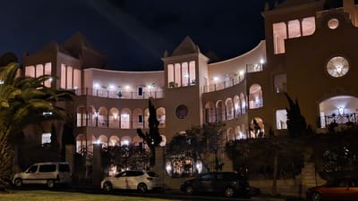 The image shows a multi-story building in Costa Adeje-playas Del Duque, Spain, taken at night. The building has a curved facade with arches, balconies, and lighted windows. Palm trees and various shrubs are in front of the building. Four cars of different makes and models are parked on the street. The sky is dark. The scene is illuminated by building lights and car headlights.