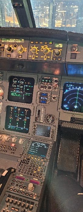 The image captures the interior of an aircraft cockpit, specifically an Airbus A320 family variant, located in Mierzęcice, Poland, consistent with Katowice Airport (KTW). The scene is set in low-light conditions, likely nighttime, indicated by the illuminated instruments and external visibility through the windshield.
No individuals are present within the frame. The primary subjects are the illuminated flight deck instruments and controls. The aircraft's windshield is visible at the top, showing reflections and apparent water droplets, with blurred external lights suggesting an airport environment.
The instrument panel features multiple active displays: two Electronic Centralized Aircraft Monitor (ECAM) displays showing green system schematics and data, and a Navigation Display (ND) on the right showing concentric blue rings. The Autopilot/Flight Control Unit (FCU) panel is visible above the displays, featuring illuminated amber digits and controls (e.g., "04000 -2.9" and "10 18"). The central pedestal contains a Multi-Control Display Unit (MCDU) with a textual screen and a full keypad below. Numerous switches, buttons, and rotary dials are illuminated with amber and green backlighting. A panel with a carbon-fiber texture is visible to the right of the MCDU. The instruments are operational, displaying information, but no human interaction is depicted.