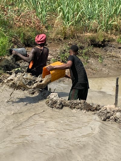 Two men work in a muddy area near a body of water. One man, wearing a maroon hat and a dark shirt, fills a black bucket with mud. The second man, in a black shirt and cap, empties a yellow container into the water. Tall green grasses line the edge. The water appears murky and disturbed by their activity. Mud and debris are caked on the embankment. A wooden post rises from the center of the water.