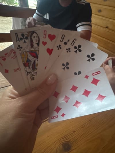 The image shows a close-up of a person's hands holding a fan of playing cards. The cards are spread out, revealing their faces.

Subjects:
*   **People:** At least one person's hands are visible, holding the cards. A portion of another person's arm and shoulder, wearing a dark shirt with white stripes, is also visible in the background.
*   **Objects:** Playing cards are the primary objects. Several card suits and numbers are visible, including diamonds, clubs, hearts, and what appear to be numbers and a queen.
*   **Scene/Setting:** The background consists of wooden planks, suggesting a rustic or indoor setting. The lighting appears to be natural, possibly daylight, casting some shadows.

Activity/Event:
The image captures the moment of someone holding or displaying a hand of playing cards, likely during a game.

Notable Details:
*   The cards are fanned out in a way that partially obscures each other.
*   Visible card values and suits include an "8 of Diamonds", "9 of Clubs", and a "Queen of Hearts" or "Queen of Clubs" with an intricate drawing. Another card appears to be a "9 of Hearts".
*   The person's fingernails are painted a light color.
*   The wooden surface has a visible grain and texture.

Visible Text:
While the card faces are partially visible, the only clearly discernible text is the number "8" on one of the diamond cards. The suits and numbers on other cards are present but partially obscured or angled in a way that makes them harder to read definitively.