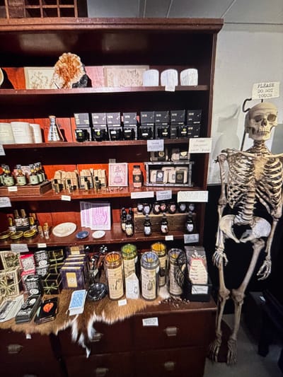 A dark wooden cabinet filled with curiosities. Shelves display small black boxes, antique-looking bottles, sealed jars with labels, and assorted trinkets. A skeleton hangs above the cabinet. A fur pelt is visible on the right side. A “Please Do Not Touch” sign is attached above the cabinet.