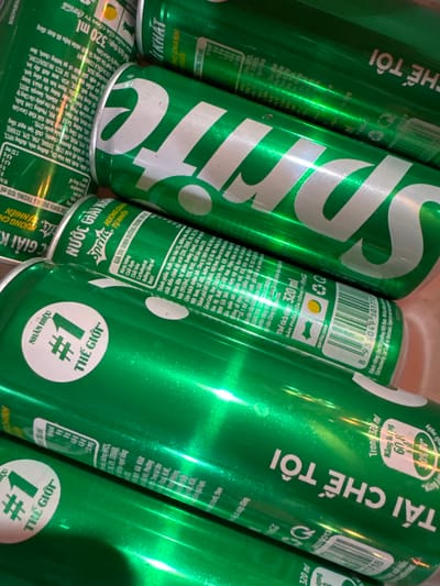 The image shows a close-up of several cans of Sprite soda. The cans are green with white lettering. Some cans display the barcode and nutritional information in Vietnamese. A round sticker on some cans says "#1 Thế giới" (World's #1 Brand). The cans are positioned close together with some cans overlapping others. No people, actions, or locations (including the specified Mèo Vạc Commune, Vietnam) are visible in this specific image. The environment appears to be indoors, with the cans possibly stored in a container or placed on a surface.