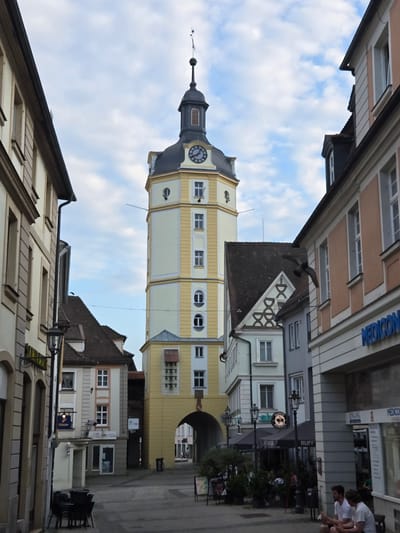 The image shows a view up a narrow European street with historical architecture. The scene is dominated by buildings, including a prominent clock tower with a yellow facade. The sky is visible with scattered clouds, suggesting daytime. Several people are present, some on the street and others in the upper floors of buildings, appearing to be engaged in everyday activities.
In the foreground, a pigeon is captured mid-flight. The buildings have varied architectural styles, with a mix of pastel and earth tones, and many windows. The clock tower has a dark spire topped with a weather vane. Visible text includes "MEDICONSULT" and "BAUER" on signs. The overall impression is of a charming, old-world European town. The angle of the shot suggests it was taken from a low vantage point, looking upwards. The lighting indicates it is likely midday or early afternoon.