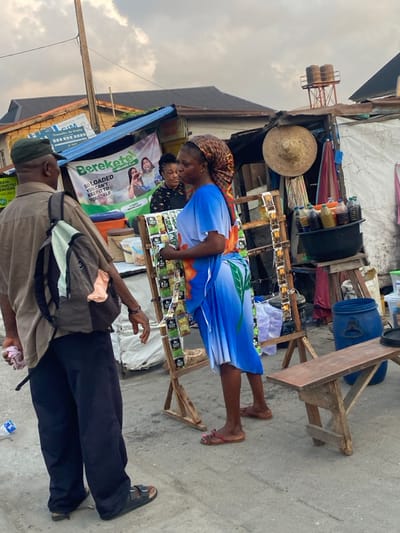A street vendor with a patterned headscarf and a blue dress stands at a wooden stall selling small packages. Behind her, a banner promoting a product is visible. A man in dark pants and a light shirt walks past, wearing sandals and carrying a backpack. The stall displays various items, including bottles of oil and a straw hat. A blue plastic barrel sits on a wooden structure nearby. The scene is set outdoors, with a building visible on the left.