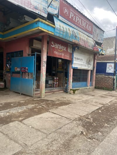 The image shows a storefront in Baleendah, Indonesia. The storefront is a single-story building with a red brick facade. There is a large, blue metal gate in front of the store, which is partially open. The storefront is adorned with various signage, including "Pi-Pamas" and "Sanlex." The storefront also displays a variety of goods, including paint cans and other building materials. The storefront appears to be located on a narrow street. The sidewalk in front of the store is cracked and uneven. The ground is covered with dirt and debris. The sky is overcast. There are no people visible in the image.