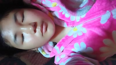 A young girl sleeps in a pink and white floral onesie. Her eyes are closed, and her head is tilted to the side, resting on a patterned blanket. The onesie is soft and fuzzy, with large daisy-like flowers printed on it. A zipper runs up the front of the onesie. The background is dark and indistinct. The image appears to be a personal photograph, likely taken in a home environment. The specific location in the city of Uchkun, Kyrgyzstan cannot be determined from the visual information present in the image.