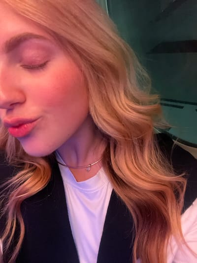 A close-up, angled view of a person lying down. The person has fair skin, closed eyes, and light pink lipstick. Their blonde hair is styled in loose waves. They are wearing a white shirt and a thin gold necklace with a teardrop pendant. The background is partially visible, with what appears to be a dark green or black surface in the upper left and a black surface to the right. No definitive details relate to the location in the city of Kempton Park, South Africa.