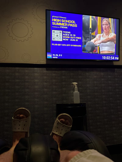 The image captures a dimly lit room, possibly a gym lounge area, with a large screen displaying an advertisement for Planet Fitness's "High School Summer Pass." The screen shows a young woman performing a kettlebell exercise. Below the screen, a spray bottle and a small table are visible. The foreground shows the lower legs and feet of one or more individuals wearing white slides, resting on black seating. The walls are textured with a subtle gear pattern. The time displayed on the screen is 10:02:54 PM. Based on the advertisements and typical gym environments, this scene is likely taking place in Tucson, Arizona, United States of America, a common location for such fitness facilities.