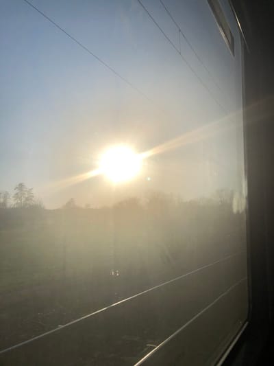 The image displays a bright, hazy, rural landscape near Dettwiller, France, viewed from the interior of a train. A low-angle, intensely radiant sun occupies the upper-left sky, producing significant lens flares and widespread atmospheric haze across the midground. Two parallel railway tracks are visible in the lower foreground, extending diagonally towards the right. Multiple thin overhead electrification wires stretch horizontally across the upper portion of the frame. Beyond the tracks, a field of vegetation is present, obscured by the glare and haze, leading to a distant line of bare deciduous trees. The right edge of the frame shows the dark structural reflection of the train's interior on the window. No individuals, specific actions, or interactions are depicted.