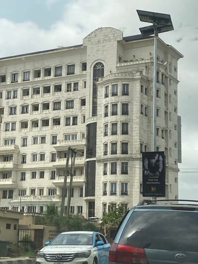 The image captures a street scene in Falomo, Nigeria, under a partially cloudy sky. A large, multi-story building with a light-colored, possibly stone, facade dominates the background. The building exhibits classical architectural features, including numerous rectangular windows, recessed balconies, a prominent central arched glass section, and decorative mouldings. Some sections of the building, particularly towards the top, show signs of surface discoloration or weathering.

In the mid-ground, a tall metal pole stands vertically, topped with a dual-panel solar street light. A dark, vertical banner is attached to the pole, displaying white text that reads "CRAFTED FOR THE DISCERNING" above a monochrome image depicting hands or an abstract form, with a web address at the bottom. To the left of this pole, a utility pole with multiple dark overhead cables is visible.

In the foreground, a dark-colored SUV is partially visible from the rear, positioned on a paved road. Its taillights and a portion of its roof rack are discernible. Directly in front of the SUV is a white sedan with blue accents on its lower body and side mirrors, also oriented to the right. Green foliage and low-lying structures are present between the road and the main building. No individuals are explicitly visible.