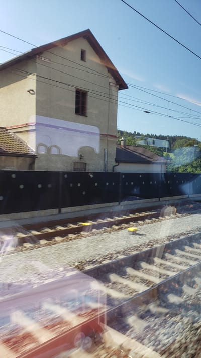 This image, captured from a moving train, shows a view of a building and train tracks. The building, a light-colored structure with a brown roof and trim, features a prominent gable and several windows. In the foreground, parallel train tracks covered in ballast are visible. A dark fence with circular cutouts separates the tracks from the area behind it, where another building with a tiled roof and a chimney is situated. Lush green trees form a backdrop to the scene. Reflections from the train window are also present, obscuring parts of the view. The location is identified as Predmier, Slovakia.