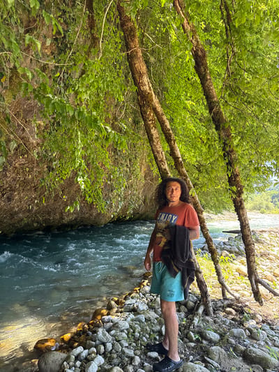 The media file depicts a person standing by a river in a natural setting. The person, with long, curly dark hair and a light complexion, is wearing a reddish-orange t-shirt with a blue graphic that reads "ENJOY Adventure" and light blue swim trunks. They are also wearing black slip-on shoes. A dark garment, possibly a jacket or towel, is draped over their shoulders.
The scene is a forest with lush green foliage and moss-covered trees. The river is on the right side of the frame, flowing with clear, turquoise water. The riverbank is lined with various-sized rocks and pebbles, some of which are illuminated by sunlight, suggesting it is daytime. The trees appear to be growing at an angle, with branches reaching out over the river and the bank.
The orientation of the image is portrait, with the river and the person appearing to be on a vertical plane. The person is looking towards the viewer with a relaxed expression. The overall impression is one of a peaceful, outdoor environment, possibly a recreational area for swimming or hiking. There are no other people visible in the frame. The location is identified as Abgara, Abkhazia. The lighting suggests it is a sunny day, and the abundance of green leaves indicates a warm season, likely spring or summer.