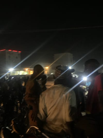At night, several people stand near a group of parked motorcycles. The lighting is poor, with bright glare spots. Some individuals wear masks. One person has on a white shirt. A tall building with red accents is visible in the distance. Power lines are visible on the left of the frame. The overall scene is cluttered and unclear due to low light and camera angle.
