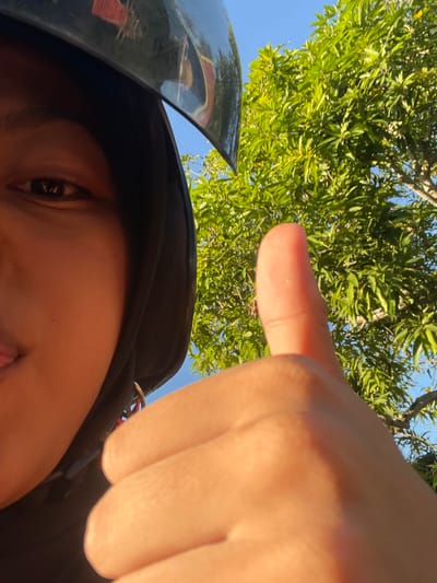This is a selfie taken from a low angle, looking up towards a person's face and a hand. The person is wearing a black helmet, with the visor reflecting a leafy tree and a blue sky. Sunlight is illuminating the scene, suggesting it is daytime. The person's face is partially visible, with one eye clearly seen. A hand is prominently featured in the foreground, with the index finger pointing upwards towards the tree. The background consists of lush green foliage and a clear blue sky, indicating an outdoor setting, likely in a tropical or sub-tropical environment. The overall impression is of a casual, personal snapshot taken on a sunny day.
