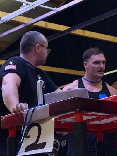 In an indoor venue in Reisenberg, Austria, two adult males are positioned around an arm wrestling table. The male on the left, with short dark hair and glasses, wears a black short-sleeved t-shirt displaying a 'HΨ' logo on the left sleeve and a blue lanyard. His left hand rests on the table's padded edge, and he is looking towards the right. The male on the right, exhibiting a muscular build, short dark hair, and a mustache, wears a black tank top with a blue rectangular patch on his right chest and a blue lanyard. His right forearm rests on a grey pad atop the table. The arm wrestling table features red and white padded perimeter, a silver vertical grip handle on the left side, and a white panel with a large black numeral '2' affixed to its front, partially covered by a blue strap. The background is dark, with visible overhead wooden beams and metallic structural supports.