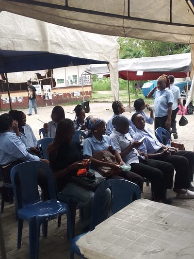 Location: Ijanikin, Nigeria.
A group of approximately 12 people, predominantly women, are seated in blue plastic chairs under a makeshift tent or awning. Most are wearing light blue shirts, some with black pants or jeans. Some individuals are using mobile phones or holding items like a small fan. A few people are standing nearby, including a woman in a light blue shirt who appears to be talking to someone.
The setting is outdoors, with a building in the background displaying posters and signage. Another tent is visible further in the background. A man is walking away in the distance near the building. A plastic table is visible in the foreground.
