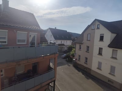 The image depicts a residential street in Kirchheim am Neckar, Germany, under a partly cloudy sky with bright sunlight. In the foreground, a multi-story building with a reddish-pink facade and balconies faces the street. The balconies have glass and metal railings, and one appears to be furnished with outdoor seating and storage. Adjacent to this building, to the right, is a pale yellow and beige building with multiple windows, some covered by rolling shutters. Further down the street, a white building with a dark roof is visible, with other residential structures and trees forming the backdrop. The street itself is paved and appears to be a quiet residential area. Two cars are parked on the street, one a silver sedan and another darker vehicle. A traffic sign with a red circle and a diagonal line is visible on a pole. The scene suggests a typical afternoon in a German town.