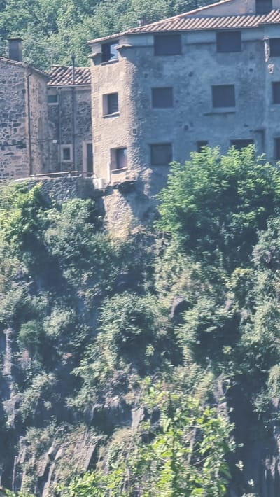 The image shows a view of the buildings of Castellfollit de la Roca, a municipality in the comarca of Garrotxa in Girona, Catalonia, Spain. The town is perched atop a basalt cliff, formed by ancient volcanic eruptions. The buildings appear to be constructed from local stone and have tile roofs. Some of the buildings are partially obscured by dense foliage, indicating a natural, hilly environment surrounding the town. The overall impression is one of a historic village integrated into a dramatic natural landscape.