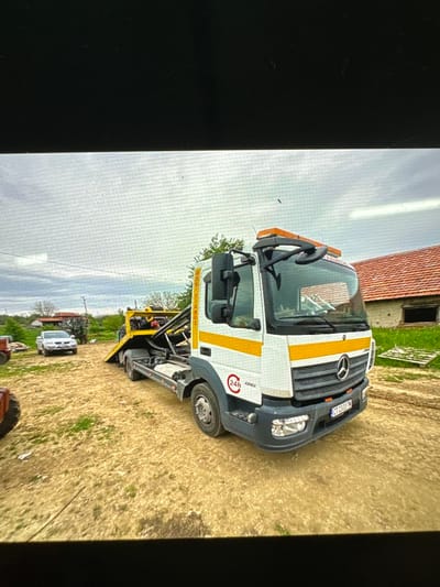This daytime image, taken in Chirpan, Bulgaria, features a white and yellow Mercedes-Benz tow truck (recovery vehicle) parked outdoors in a rural or semi-rural setting. The truck is positioned on dry, yellowish-brown ground with scattered patches of green grass, under an overcast sky.
The prominent subject is the tow truck, painted white with distinctive horizontal yellow stripes on the cab's side and across the front grille. It has the Mercedes-Benz logo on its grille and appears to be a 916 model, as indicated by numbers on the cab. Orange beacon lights are mounted on its roof. The front license plate displays "CT 2337 M", with "CT" signifying Stara Zagora, the region where Chirpan is located. On the side of the cab, a red circular emblem with "24h" and an arrow indicates 24-hour service, below which a partially visible text resembling "EPS..." is present. The truck is equipped with heavy-duty recovery gear at the rear, including a flatbed and a lift mechanism, with some yellow text on black strapping visible but illegible.
In the background, a building with a red tiled roof and a dark, rectangular window opening suggests a barn or old house. Further back, another white vehicle, possibly a pickup truck or SUV, is visible, along with parts of what appears to be agricultural machinery, such as a tractor wheel. Trees and distant foliage line the horizon under a grey, cloudy sky. No people are visible in the scene, and no specific activity beyond the truck being parked is taking place. The overall impression is one of a working vehicle in an agricultural or peripheral urban environment.